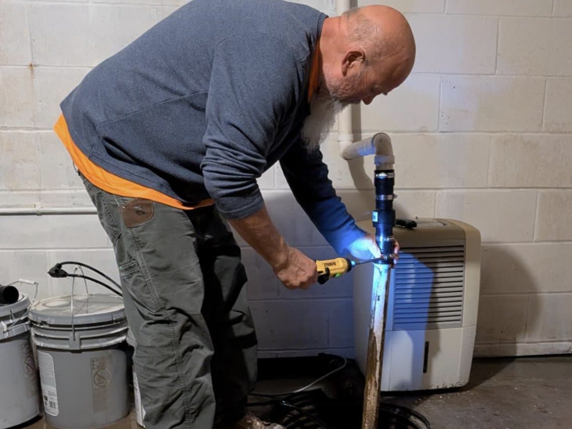 Bob bent over a basement sump pit, drill in one hand and a flashlight on the discharge pipe, tightening the fitting